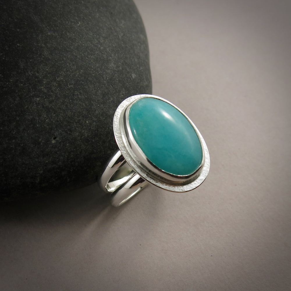A sterling silver & gemstone ring is displayed on a grey background.  The oval Amazonite gemstone is a pretty blue-green and is bezel set in silver.  The band is a split shank.