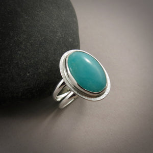 A sterling silver & gemstone ring is displayed on a grey background.  The oval Amazonite gemstone is a pretty blue-green and is bezel set in silver.  The band is a split shank.