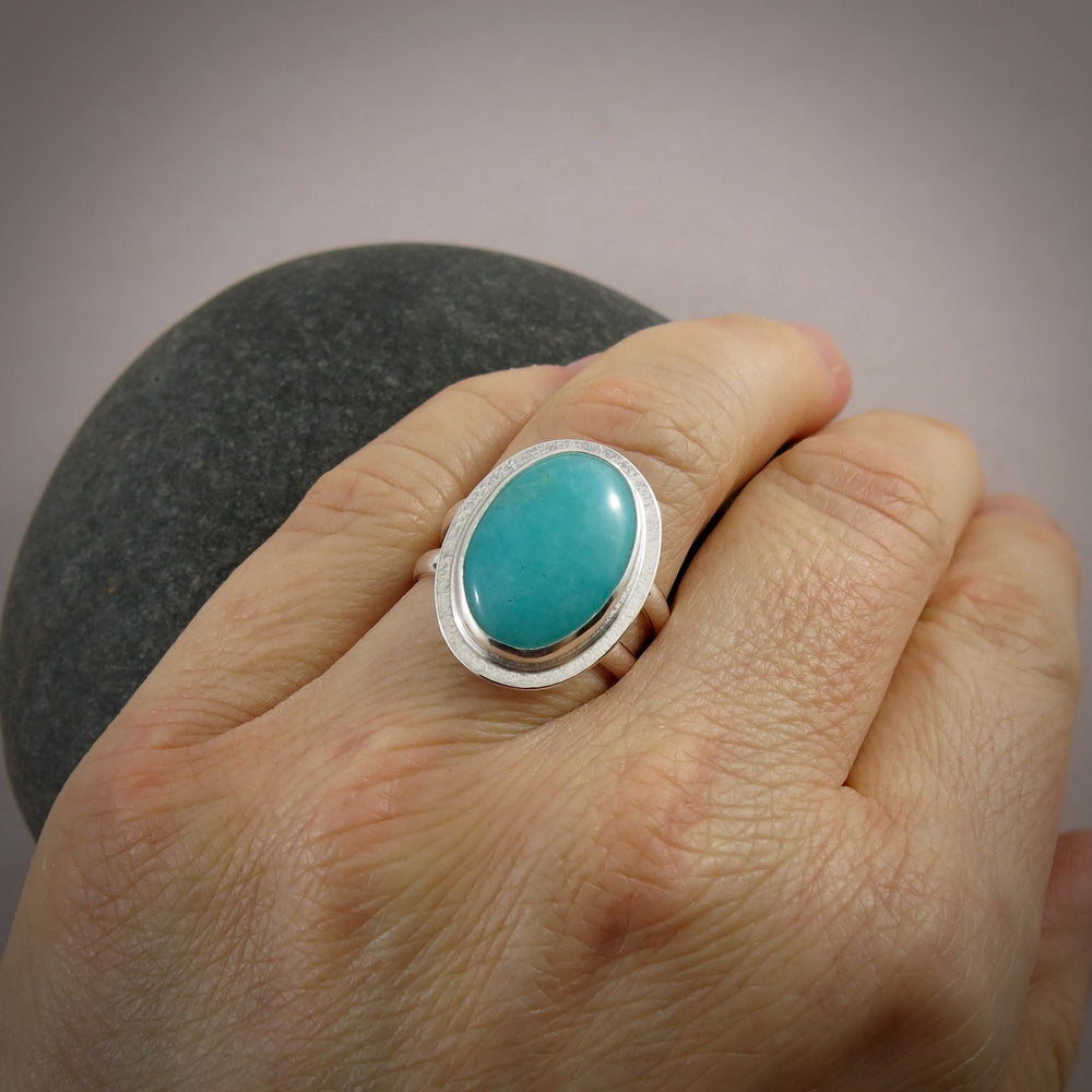 A sterling silver & gemstone ring is displayed on a hand.  The oval Amazonite gemstone is a pretty blue-green and is bezel set in silver.  The band is a split shank.