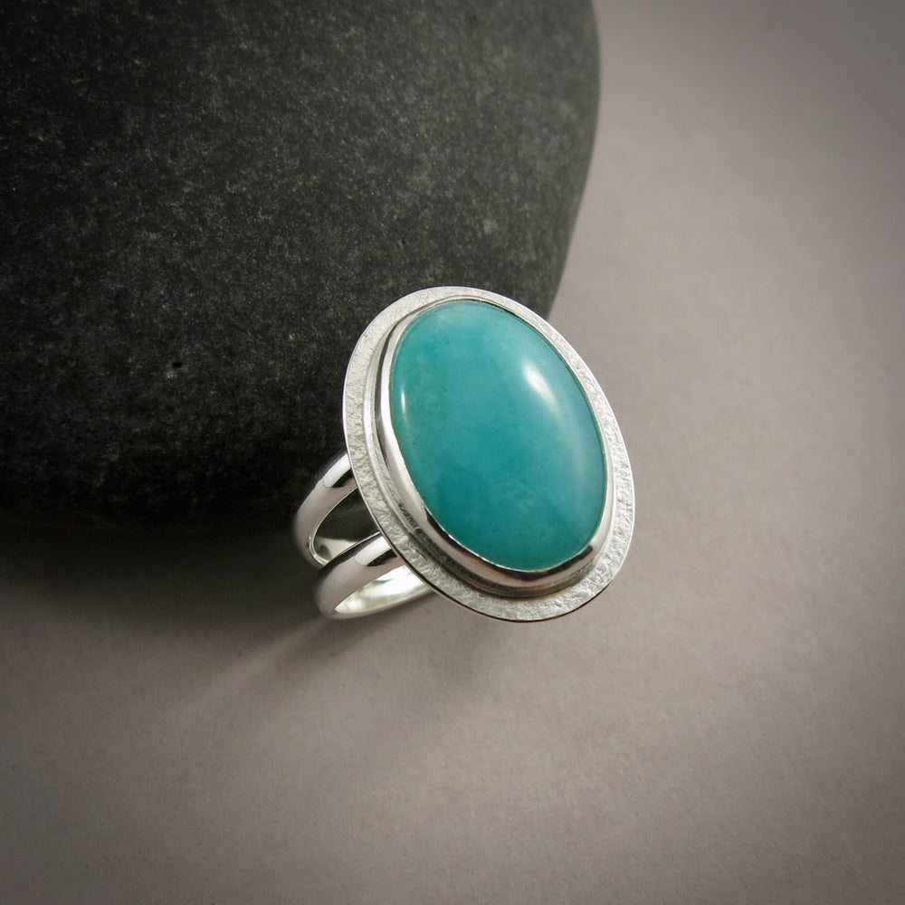 A sterling silver & gemstone ring is displayed on a grey background.  The oval Amazonite gemstone is a pretty blue-green and is bezel set in silver.  The band is a split shank.