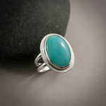 A sterling silver & gemstone ring is displayed on a grey background.  The oval Amazonite gemstone is a pretty blue-green and is bezel set in silver.  The band is a split shank.