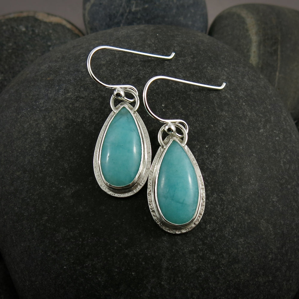 A pair of earrings is displayed on a black beach rock.  The earrings feature elongated teardrop shaped amazonite gemstones in a lovely sea foam blue-green.  The gems are bezel set in silver and have a subtly textured silver frame surrounding them.  