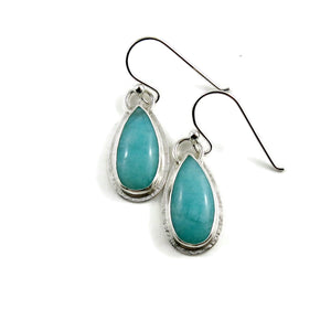 A pair of earrings is displayed on a white background.  The earrings feature elongated teardrop shaped amazonite gemstones in a lovely sea foam blue-green.  The gems are bezel set in silver and have a subtly textured silver frame surrounding them.  