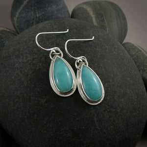 A pair of earrings is displayed on a black beach rock.  The earrings feature elongated teardrop shaped amazonite gemstones in a lovely sea foam blue-green.  The gems are bezel set in silver and have a subtly textured silver frame surrounding them.  