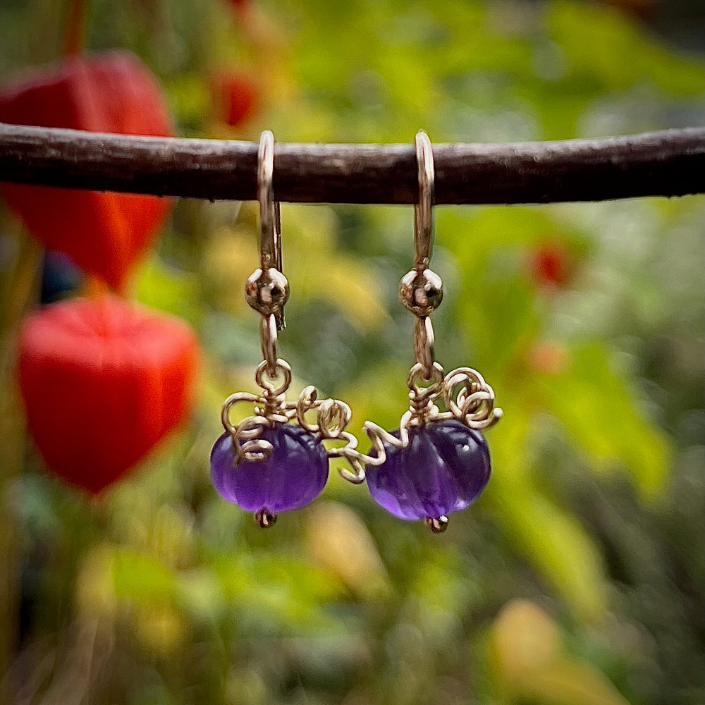 A pair of earrings are hanging on a branch with blurred natural background.  The earrings feature carved purple amethyst beads that look like tiny pumpkins with curled gold filled wire that looks like vines atop.  They hang from gold filled ear wires.