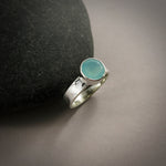 A sterling silver ring with a soft green faceted round gemstone is displayed on a dark background against a black beach rock.  The ring has a hammer textured band.