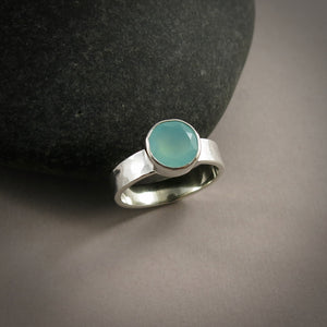 A sterling silver ring with a soft green faceted round gemstone is displayed on a dark background against a black beach rock.  The ring has a hammer textured band.
