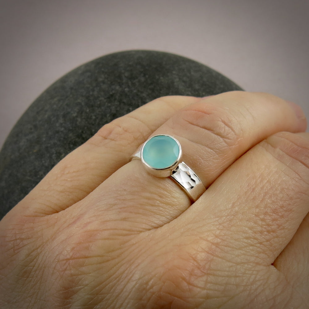 A sterling silver ring with a soft green faceted round gemstone is displayed on a hand.  The ring has a hammer textured band.