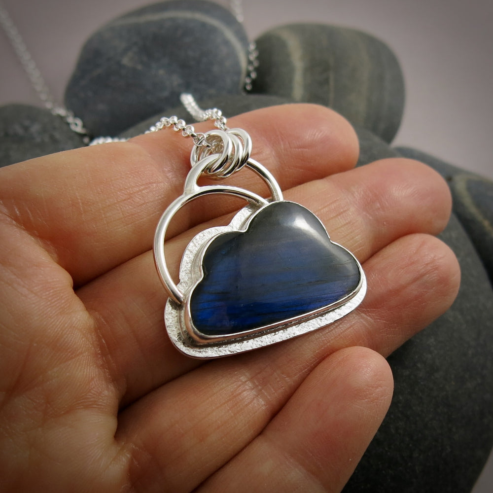 A "Silver Linings" necklace by Mikel Grant Jewellery.  An electric blue labradorite gemstone in the shape of a cloud is bezel set in sterling silver on a silver back.  A "silver lining" of silver surrounds the stone and a single silver "rainbow" sits above the cloud.
