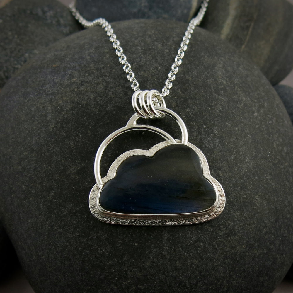 A "Silver Linings" necklace by Mikel Grant Jewellery.  An electric blue labradorite gemstone in the shape of a cloud is bezel set in sterling silver on a silver back.  A "silver lining" of silver surrounds the stone and a single silver "rainbow" sits above the cloud.