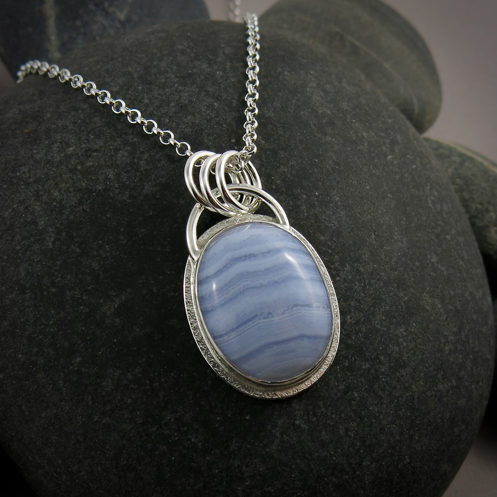A silver necklace with a gemstone pendant is displayed on a black beach rock.  The pendant features an oval, softly banded blue lace agate gemstone bezel set in silver with a subtly textured "halo" surround.