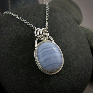 A silver necklace with a gemstone pendant is displayed on a black beach rock.  The pendant features an oval, softly banded blue lace agate gemstone bezel set in silver with a subtly textured "halo" surround.