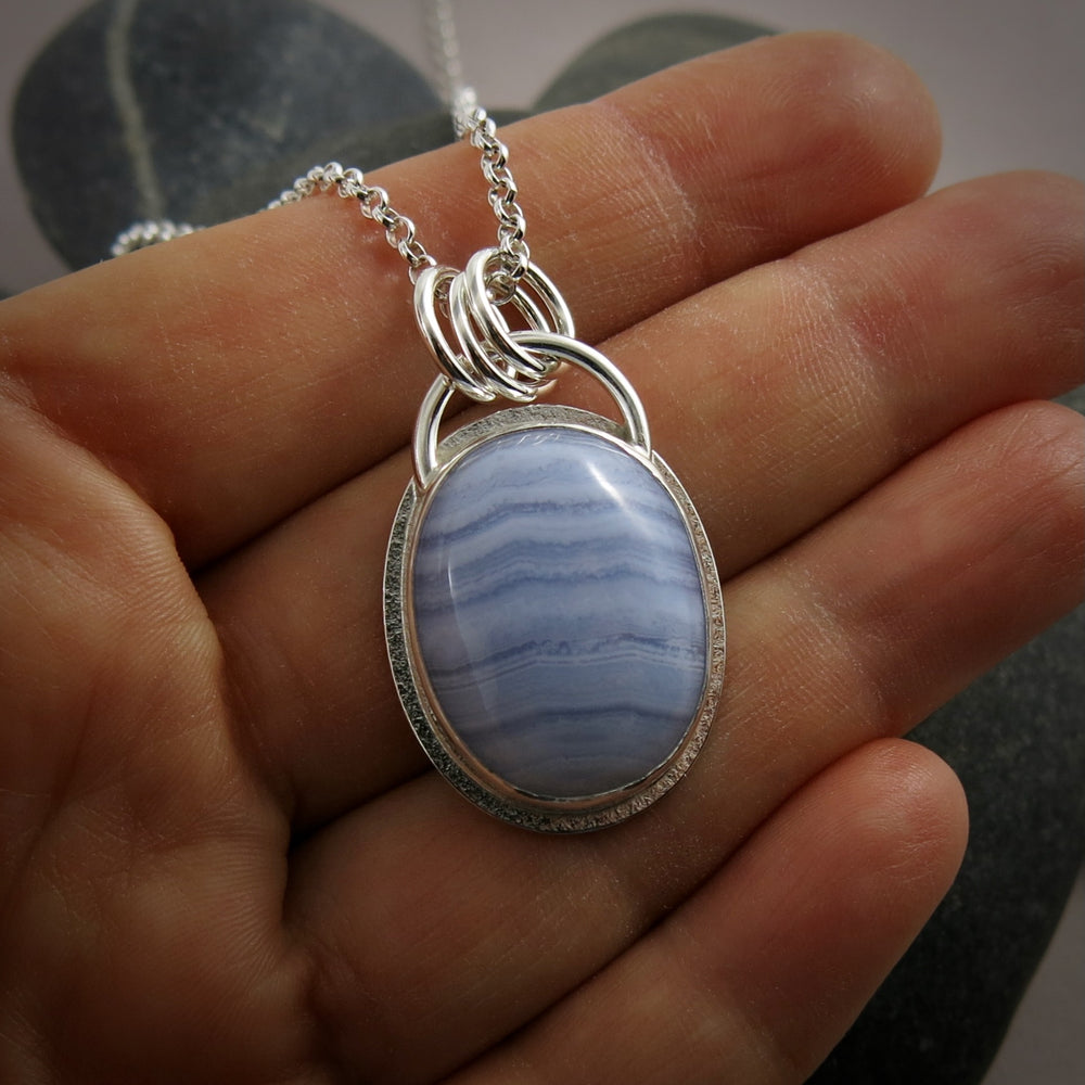 A silver necklace with a gemstone pendant is displayed on a hand.  The pendant features an oval, softly banded blue lace agate gemstone bezel set in silver with a subtly textured "halo" surround.