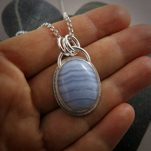 A silver necklace with a gemstone pendant is displayed on a hand.  The pendant features an oval, softly banded blue lace agate gemstone bezel set in silver with a subtly textured "halo" surround.