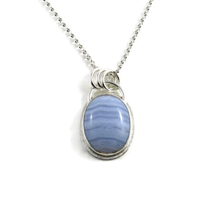 A silver necklace with a gemstone pendant is displayed on a white background.  The pendant features an oval, softly banded blue lace agate gemstone bezel set in silver with a subtly textured "halo" surround.