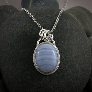 A silver necklace with a gemstone pendant is displayed on a black beach rock.  The pendant features an oval, softly banded blue lace agate gemstone bezel set in silver with a subtly textured "halo" surround.