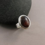 A sterling silver ring with an oval Botswana agate gemstone is displayed on a grey background.  The stone features colour bands of orange, maroon and chocolate brown.