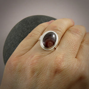 A sterling silver ring with an oval Botswana agate gemstone is displayed on a  hand.  The stone features colour bands of orange, maroon and chocolate brown.