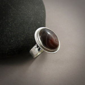 A sterling silver ring with an oval Botswana agate gemstone is displayed on a grey background.  The stone features colour bands of orange, maroon and chocolate brown.