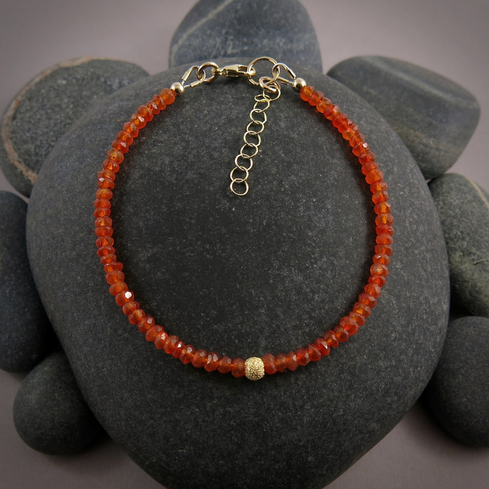 A bright orange gemstone bead bracelet is displayed on a black beach rock.  The carnelian beads are faceted and sparkly and the bracelet features a gold filled sparkly bead in the centre as well as a gold filled clasp, ring and adjustable chain.