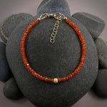 A bright orange gemstone bead bracelet is displayed on a black beach rock.  The carnelian beads are faceted and sparkly and the bracelet features a gold filled sparkly bead in the centre as well as a gold filled clasp, ring and adjustable chain.