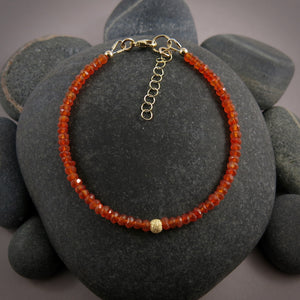 A bright orange gemstone bead bracelet is displayed on a black beach rock.  The carnelian beads are faceted and sparkly and the bracelet features a gold filled sparkly bead in the centre as well as a gold filled clasp, ring and adjustable chain.