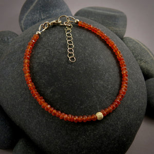 A bright orange gemstone bead bracelet is displayed on a black beach rock.  The carnelian beads are faceted and sparkly and the bracelet features a gold filled sparkly bead in the centre as well as a gold filled clasp, ring and adjustable chain.