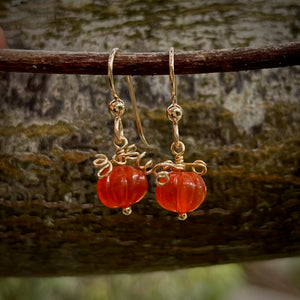 A pair of earrings are hanging on a branch with blurred natural background.  The earrings feature carved orange carnelian beads that look like tiny pumpkins with curled gold filled wire that looks like vines atop.  They hang from gold filled ear wires.