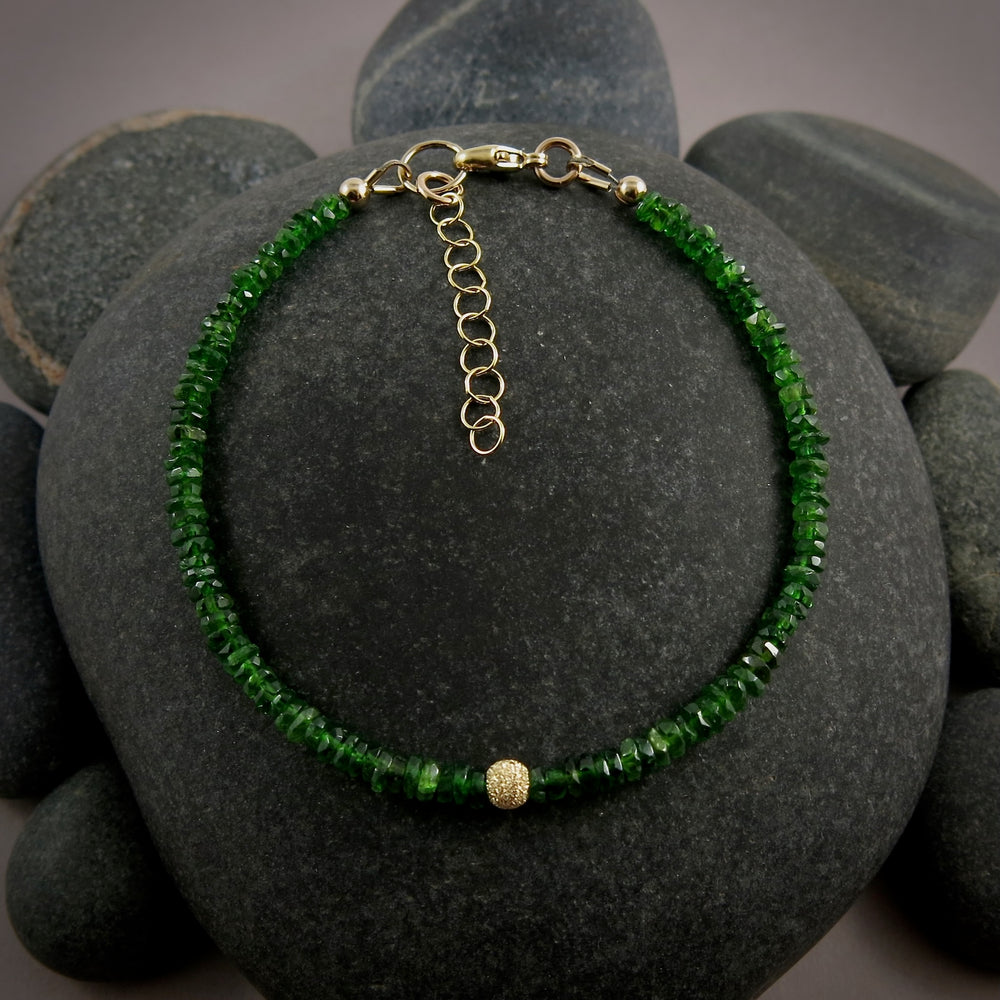 A bright green gemstone bead bracelet is displayed on a black beach rock.  The chrome diopside beads are faceted and sparkly and the bracelet features a gold filled sparkly bead in the centre as well as gold filled clasp, ring and adjustable chain.