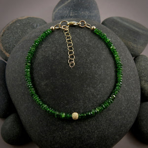 A bright green gemstone bead bracelet is displayed on a black beach rock.  The chrome diopside beads are faceted and sparkly and the bracelet features a gold filled sparkly bead in the centre as well as gold filled clasp, ring and adjustable chain.