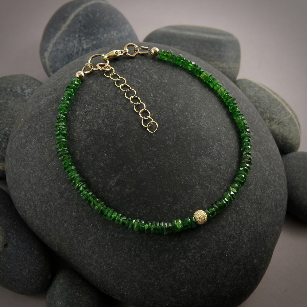 A bright green gemstone bead bracelet is displayed on a black beach rock.  The chrome diopside beads are faceted and sparkly and the bracelet features a gold filled sparkly bead in the centre as well as gold filled clasp, ring and adjustable chain.