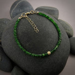 A bright green gemstone bead bracelet is displayed on a black beach rock.  The chrome diopside beads are faceted and sparkly and the bracelet features a gold filled sparkly bead in the centre as well as gold filled clasp, ring and adjustable chain.