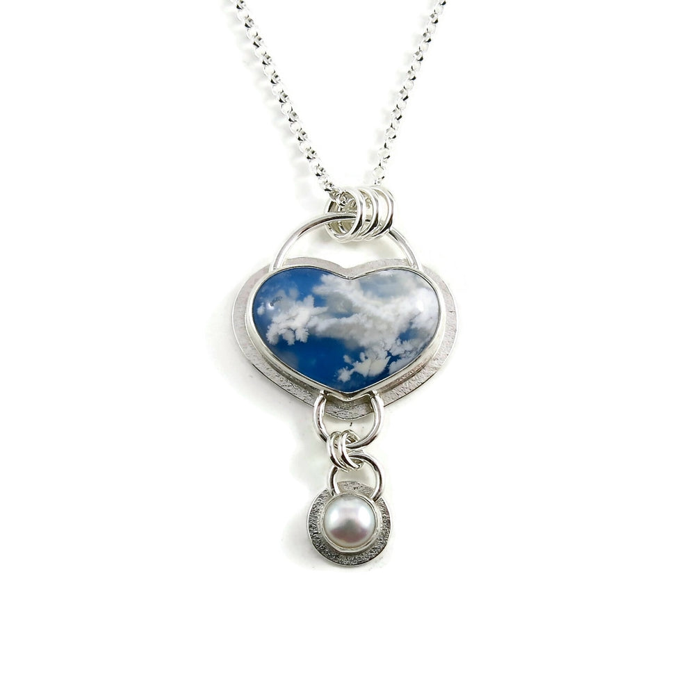 Daydream necklace by Mikel Grant Jewellery.  A heart shaped plume agate displays fluffy white clouds against a bright blue sky-looking background.   A bezel set white button pearl dangles from the heart.  