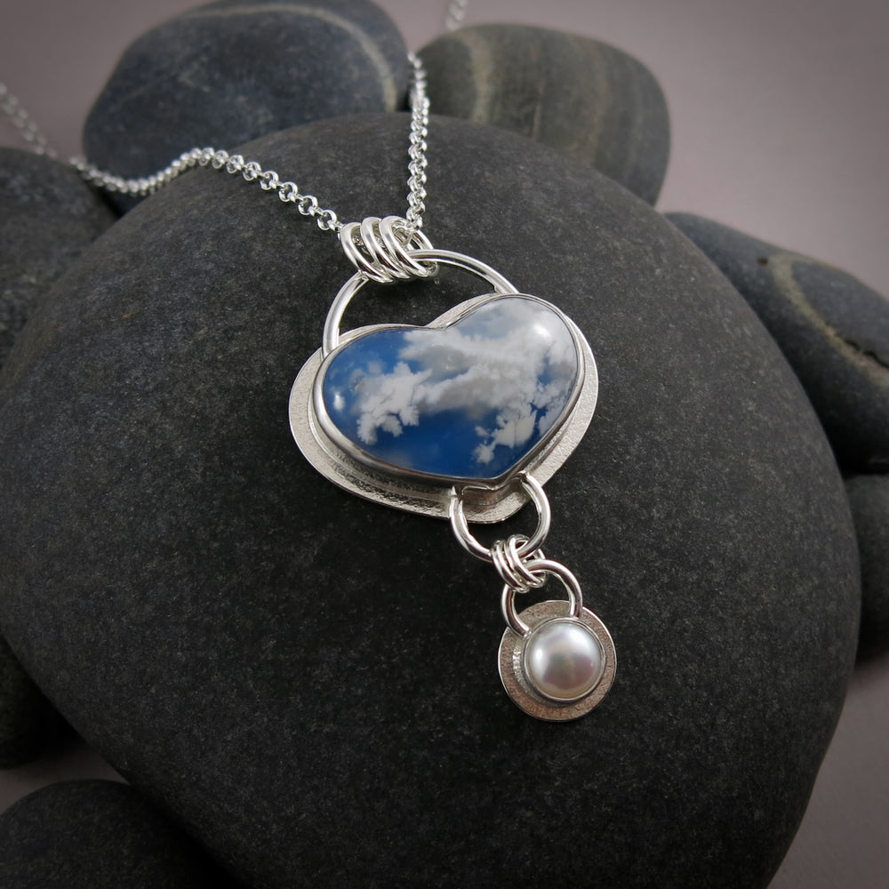 Daydream necklace by Mikel Grant Jewellery.  A heart shaped plume agate displays fluffy white clouds against a bright blue sky-looking background.   A bezel set white button pearl dangles from the heart.  