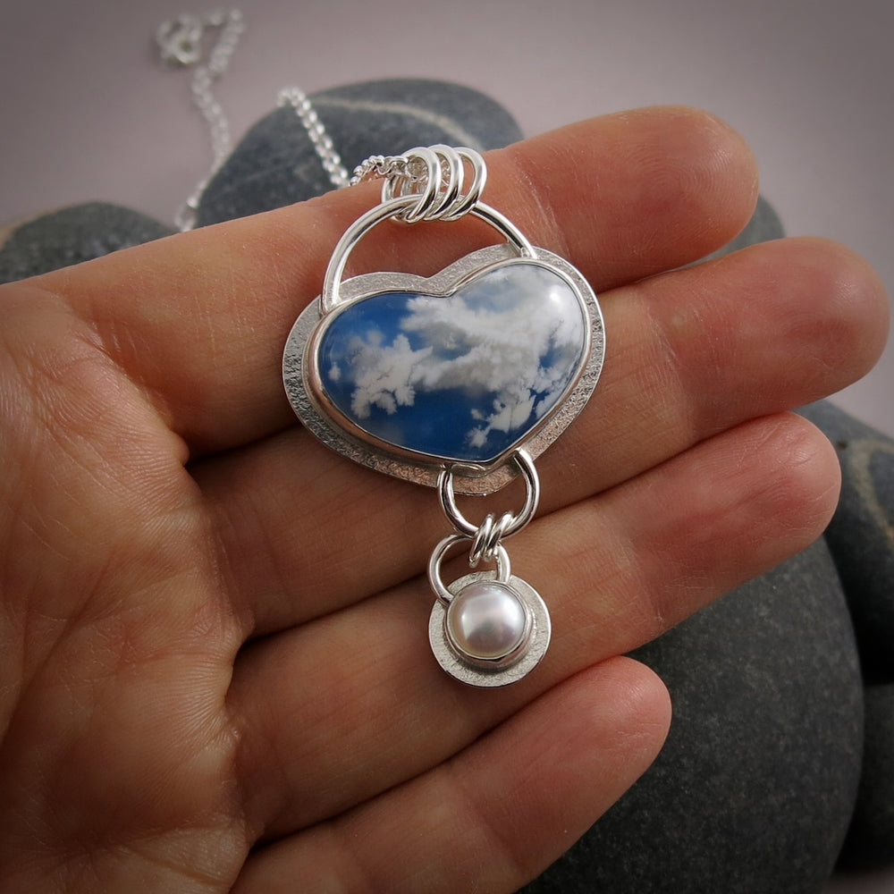 Daydream necklace by Mikel Grant Jewellery.  A heart shaped plume agate displays fluffy white clouds against a bright blue sky-looking background.   A bezel set white button pearl dangles from the heart.  