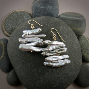 A pair of grey pearl earrings are displayed on a black beach rock.  The earrings feature a stack of five organically shaped keshi toothpick earrings on a gold headpin with gold ear wires.