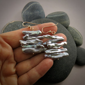 A pair of grey pearl earrings are displayed on a hand.  The earrings feature a stack of five organically shaped keshi toothpick earrings on a gold headpin with gold ear wires.