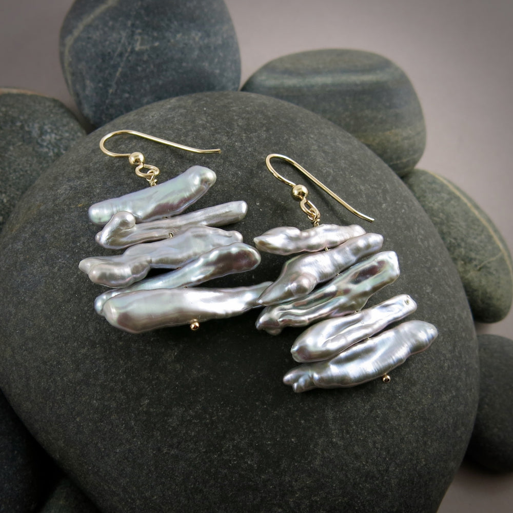 A pair of grey pearl earrings are displayed on a black beach rock.  The earrings feature a stack of five organically shaped keshi toothpick earrings on a gold headpin with gold ear wires.