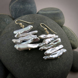 A pair of grey pearl earrings are displayed on a black beach rock.  The earrings feature a stack of five organically shaped keshi toothpick earrings on a gold headpin with gold ear wires.