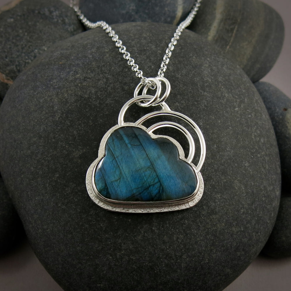 A sterling silver necklace by Mikel Grant Jewellery.  Featuring an electric blue labradorite gemstone in the shape of a cloud.   The labradorite is bezel set in silver with a silver "lining" frame around the outside and a double "rainbow" above the cloud made from two silver curved rings.