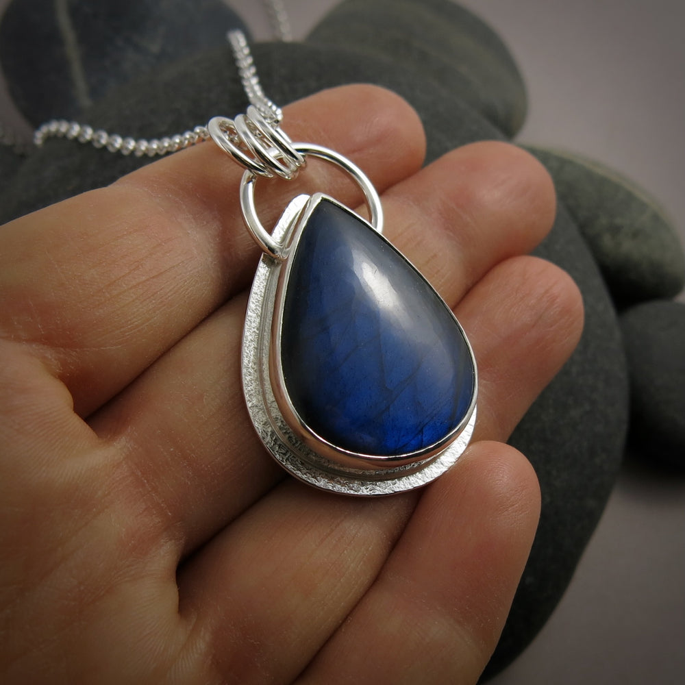 A silver necklace with a gemstone pendant is displayed on a hand.  The pendant features a teardrop shaped electric blue labradorite gemstone set in a silver bezel and a subtly textured silver "halo" surround.