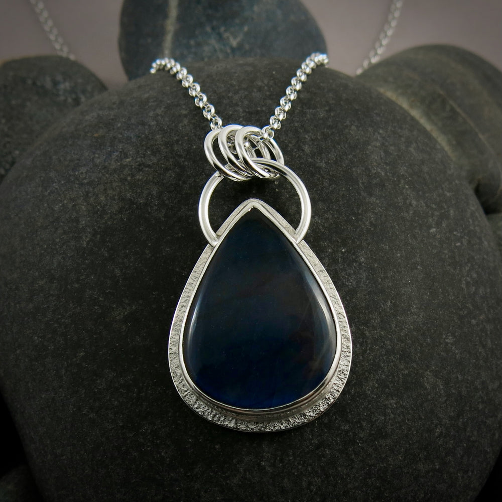 A silver necklace with a gemstone pendant is displayed on a white background.  The pendant features a teardrop shaped electric blue labradorite gemstone set in a silver bezel and a subtly textured silver "halo" surround.