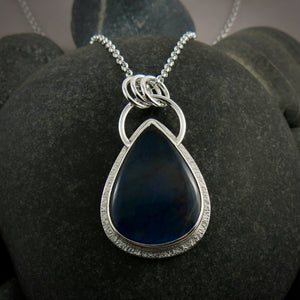 A silver necklace with a gemstone pendant is displayed on a white background.  The pendant features a teardrop shaped electric blue labradorite gemstone set in a silver bezel and a subtly textured silver "halo" surround.