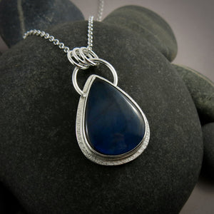 A silver necklace with a gemstone pendant is displayed on a black beach rock.  The pendant features a teardrop shaped electric blue labradorite gemstone set in a silver bezel and a subtly textured silver "halo" surround.