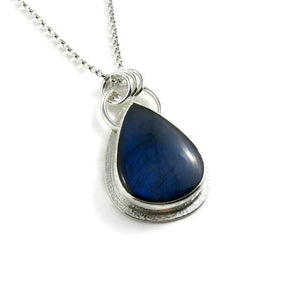 A silver necklace with a gemstone pendant is displayed on a white background.  The pendant features a teardrop shaped electric blue labradorite gemstone set in a silver bezel and a subtly textured silver "halo" surround.