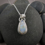 A rainbow moonstone necklace is displayed on a black beach rock.  The elongated teardrop showcases a lovely blue glow and rainbow flash across the centre of the stone.  The stone is set in sterling silver.
