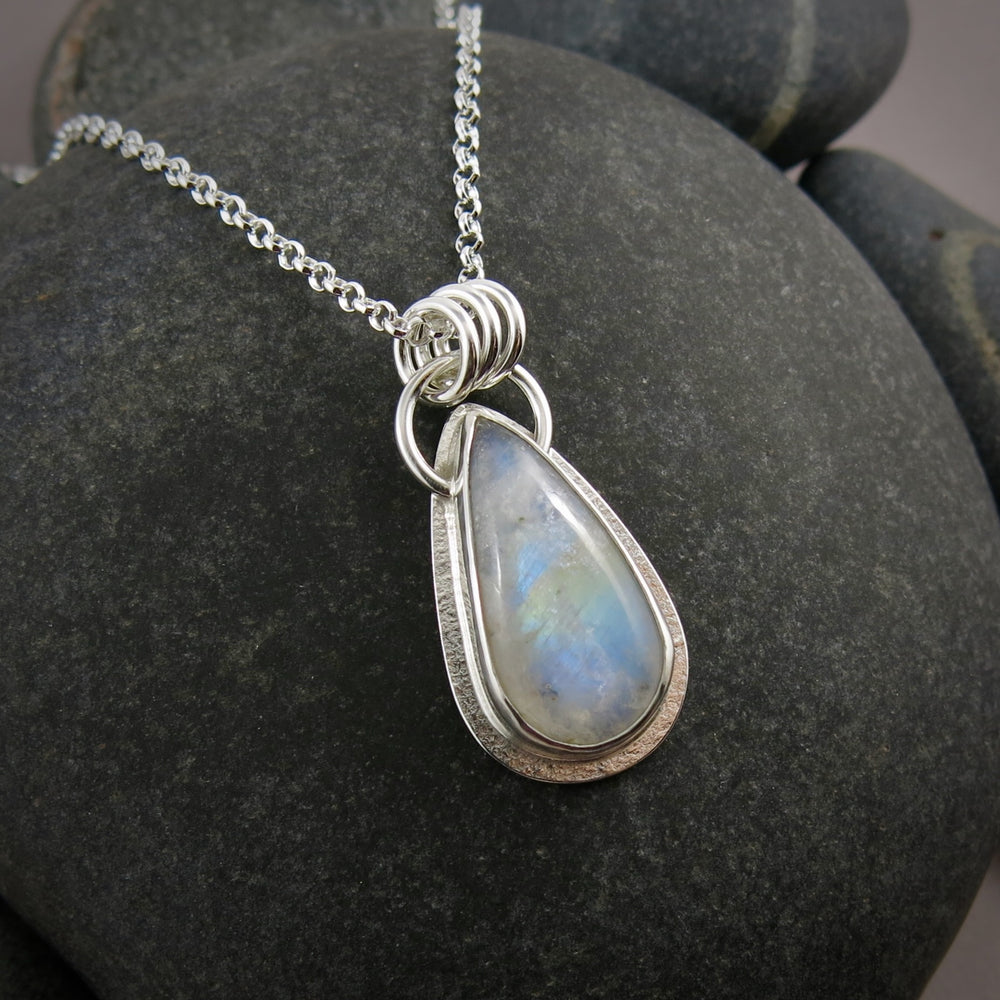 A rainbow moonstone necklace is displayed on a black beach rock.  The elongated teardrop showcases a lovely blue glow and rainbow flash across the centre of the stone.  The stone is set in sterling silver.