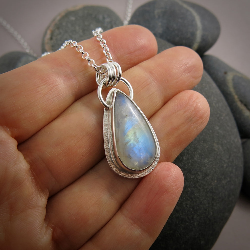 A rainbow moonstone necklace is displayed on a hand.  The elongated teardrop showcases a lovely blue glow and rainbow flash across the centre of the stone.  The stone is set in sterling silver.