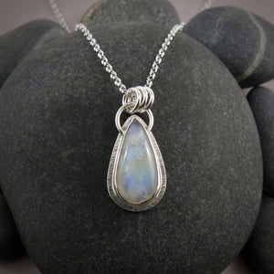 A rainbow moonstone necklace is displayed on a black beach rock.  The elongated teardrop showcases a lovely blue glow and rainbow flash across the centre of the stone.  The stone is set in sterling silver.
