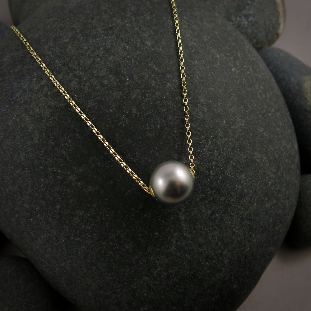 A pearl necklace is displayed on a black beach rock.  The round, gray pearl is suspended on a yellow gold filled rolo chain that slides through the centre of the pearl.